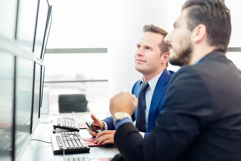 Two businessmen analyzing property investment data on multiple screens, discussing interstate investment strategies.