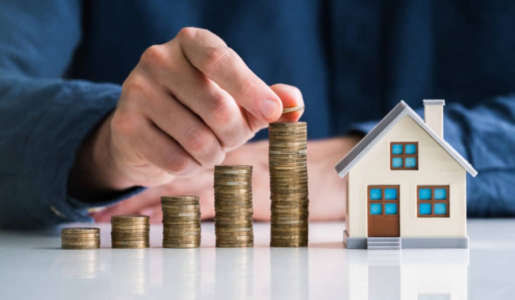 Person stacking coins next to a model house, symbolizing real estate investment growth.